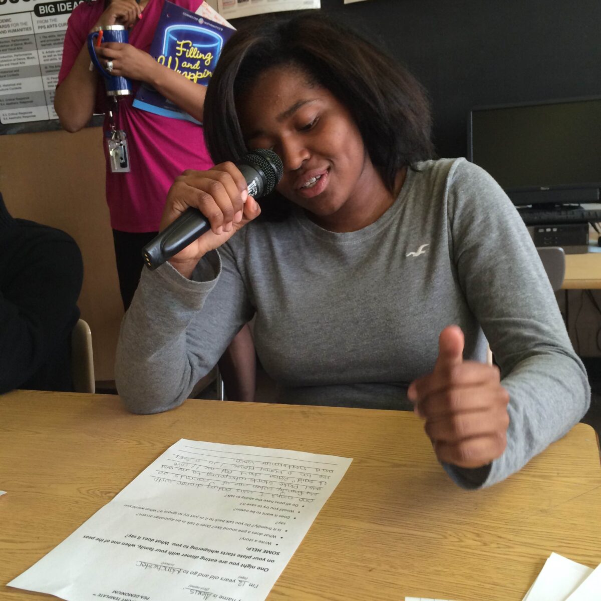 IMG_1080 A youth holding and speaking into a microphone looking down at a piece of paper on a classroom desk