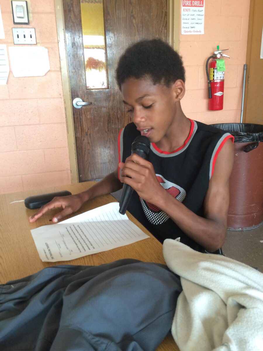 A youth holding and speaking into a microphone looking down at a piece of paper on a classroom desk