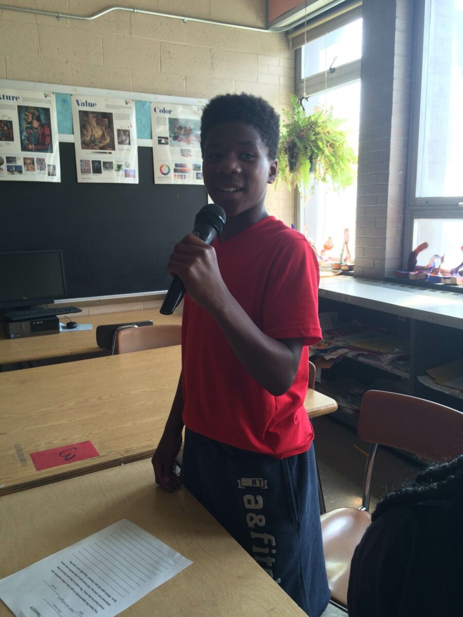 A youth holding and speaking into a microphone standing next to a classroom desk