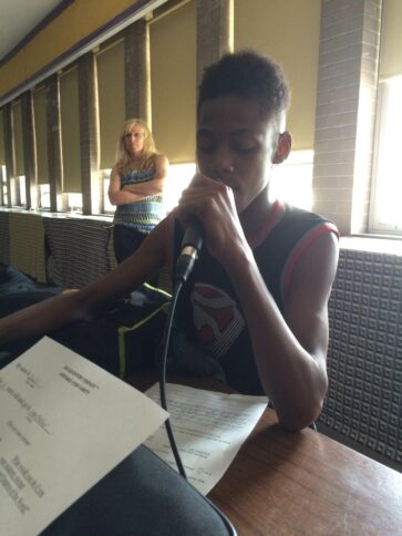 A youth holding a microphone and reading a piece of paper sitting on a classroom desk as an adult watches