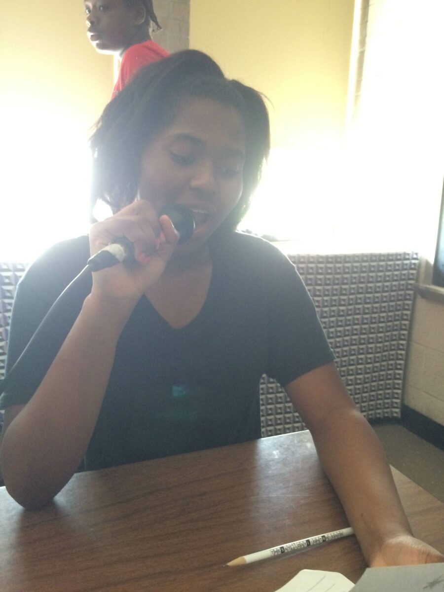 A youth holding and speaking into a microphone looking down at a piece of paper sitting at a classroom desk