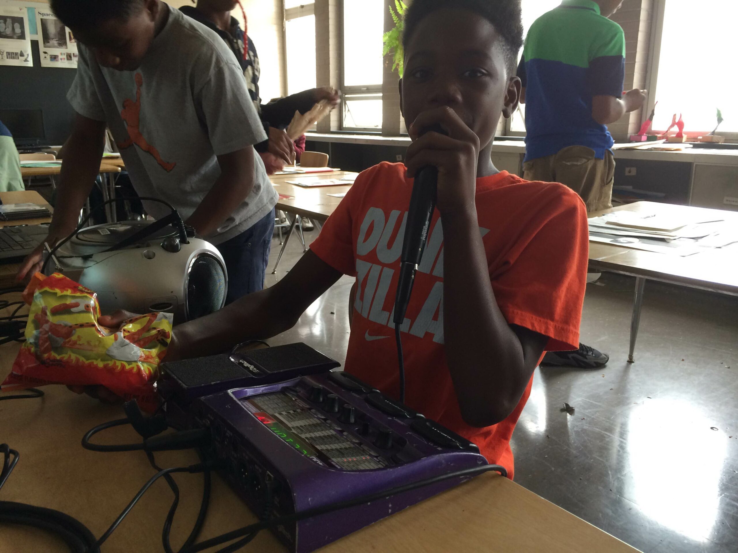 A youth holding and speaking into a microphone connected to a small mixer on a table in a classroom