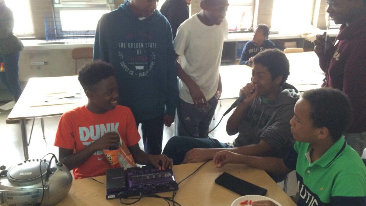 Six youth standing around a mixer and a cd player on a classroom desk, one youth holding a microphone
