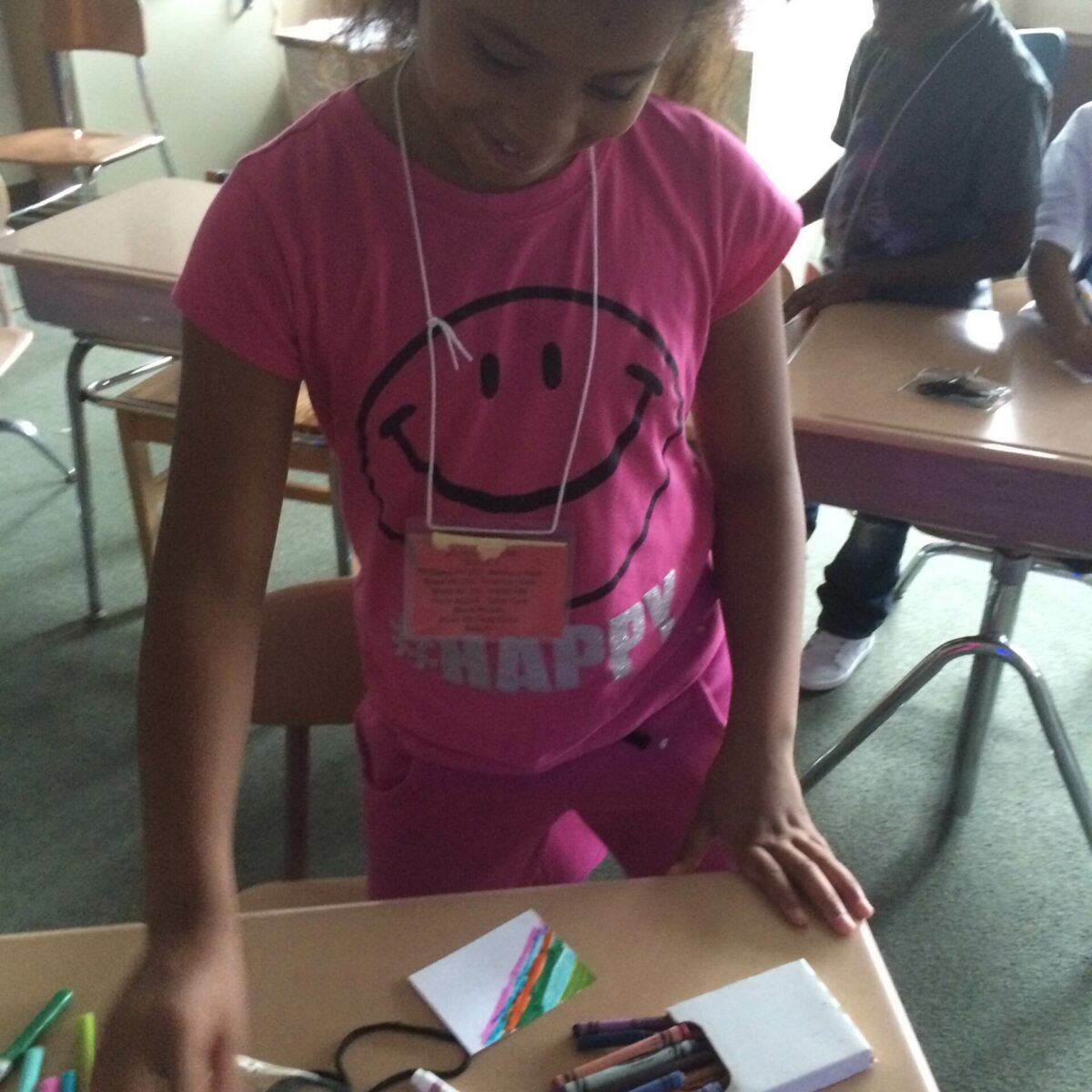 A youth smiling while grabbing markers on a classroom desk