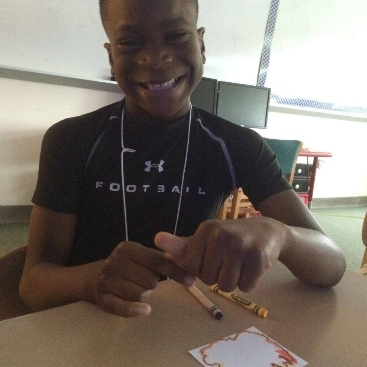 A youth smiling while sitting behind a piece of paper and two crayons on a classroom desk