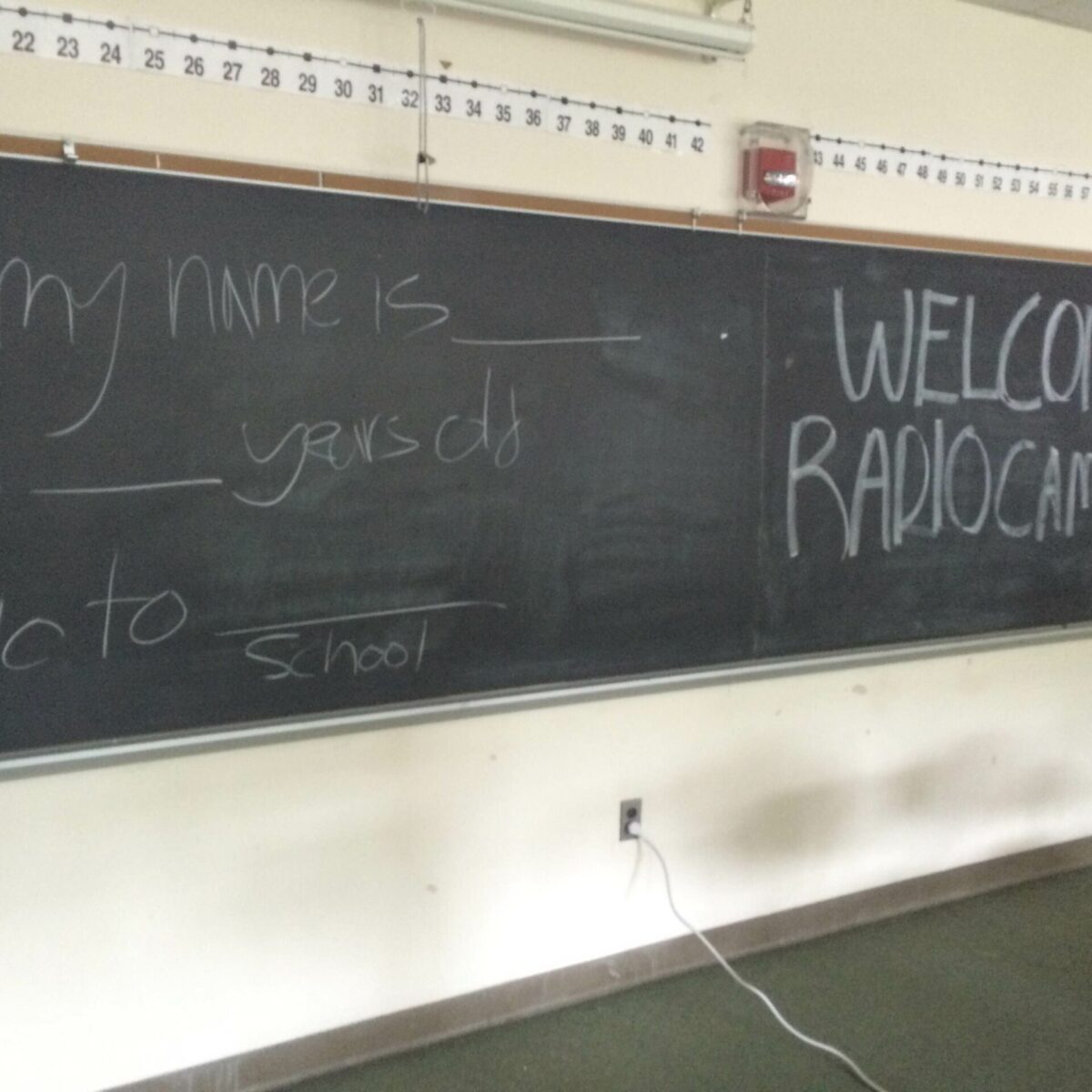 A chalkboard with the text "Hi my name is ___ I'm ___ years olds I go to ___school" on the left and "Welcome to Radio Camp!" on the right