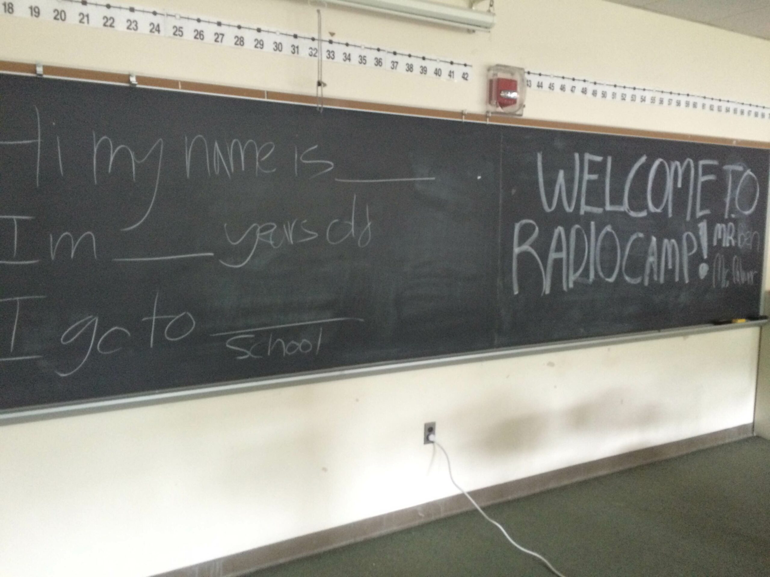 A chalkboard with the text "Hi my name is ___ I'm ___ years olds I go to ___school" on the left and "Welcome to Radio Camp!" on the right