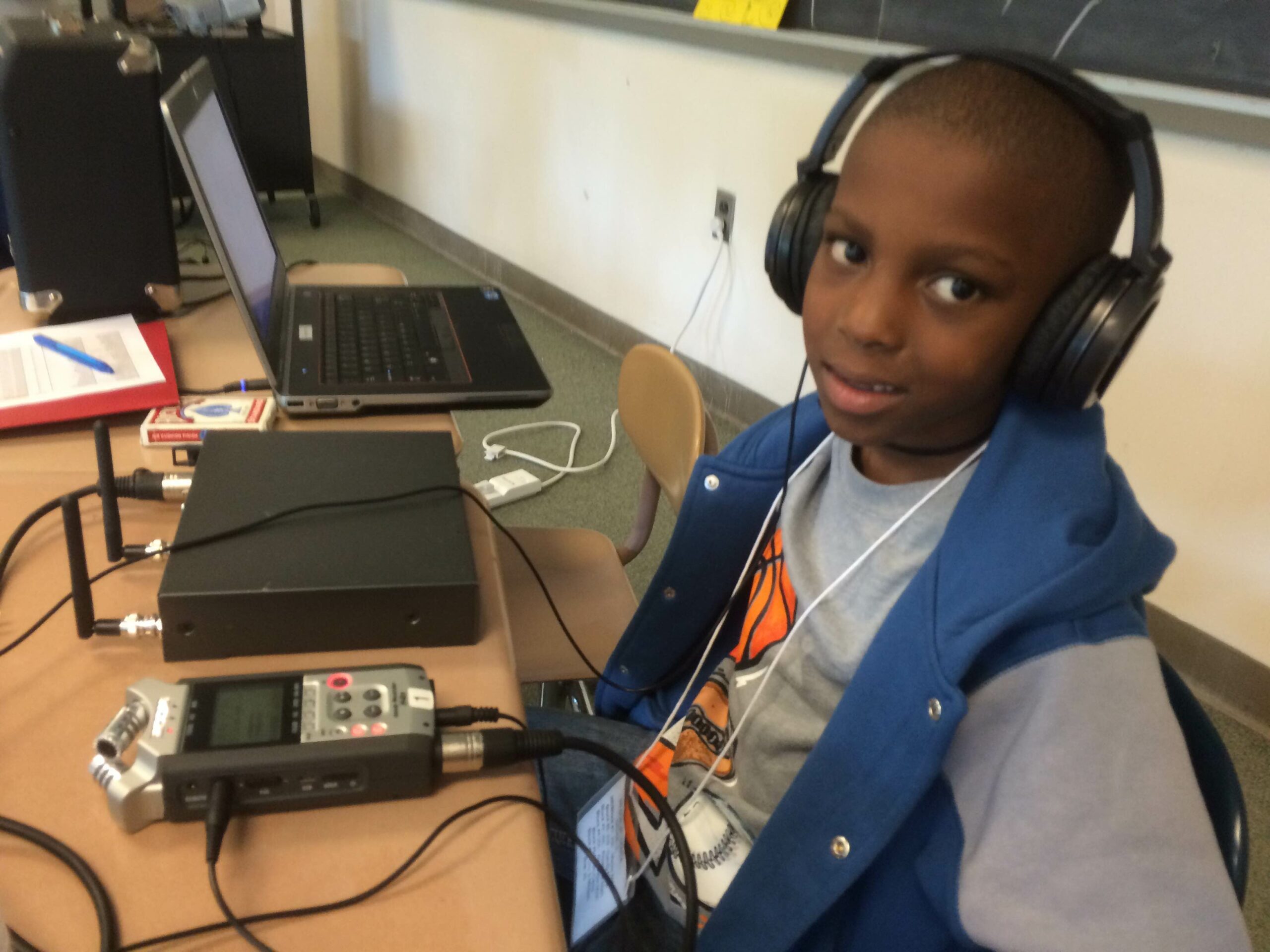 A youth wearing headphones sitting behind a portable recorder, a wireless receiver, and a laptop computer on a classroom desk