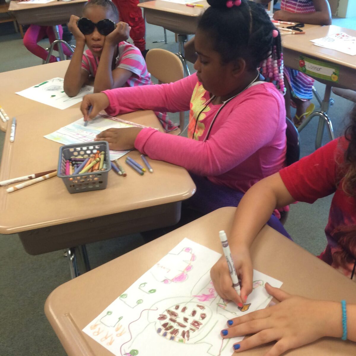 Two youth drawing on pieces of paper and one youth holding sunglasses on their face, all sitting at classroom desks