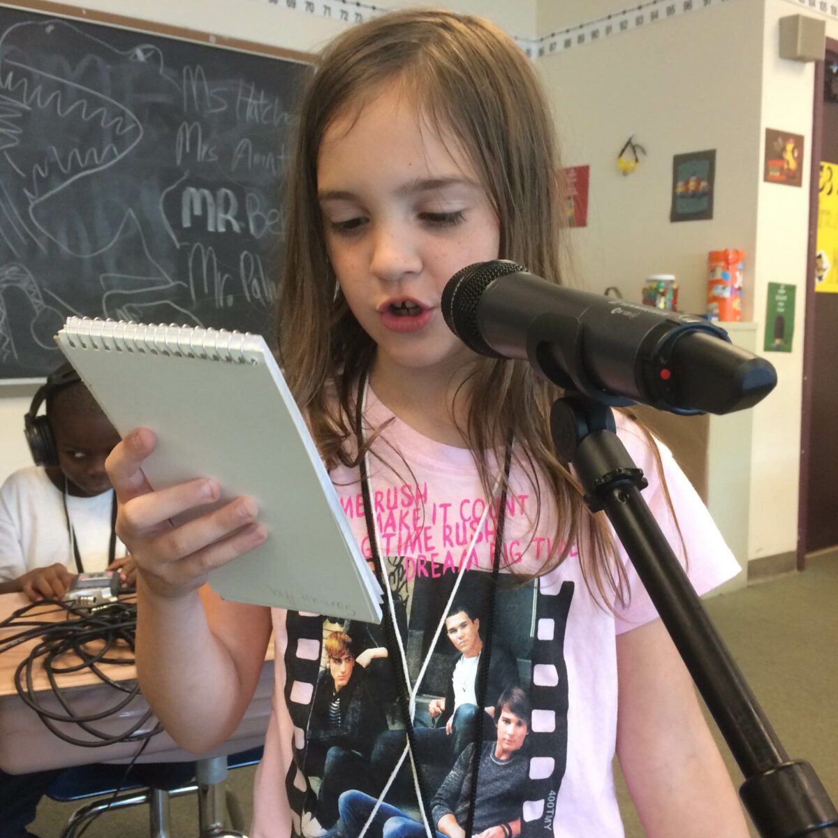 A youth looking at a notepad and speaking into a microphone in a classroom