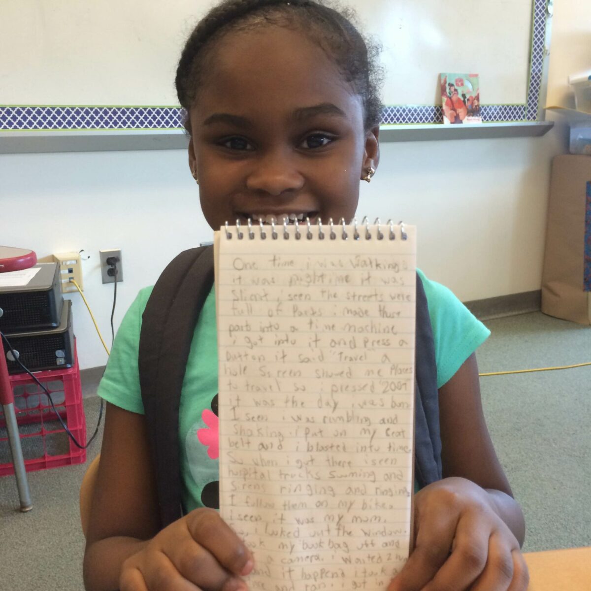 A youth smiling while holding up a notepad sitting at a classroom desk