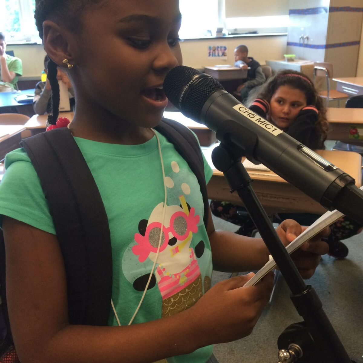 A youth holding a notepad and speaking into a microphone in a classroom