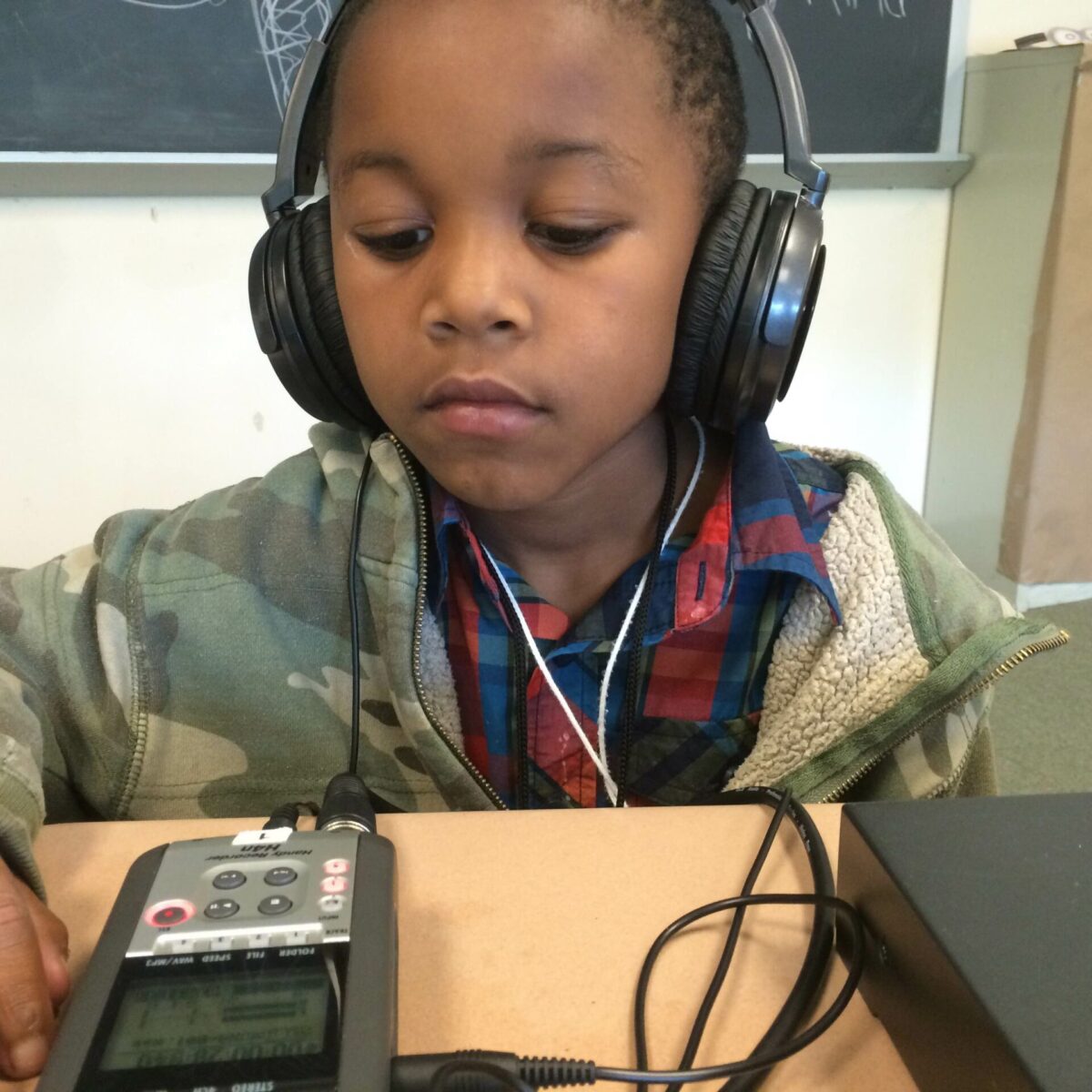 A youth wearing headphones looking down at a portable recorder on a classroom desk