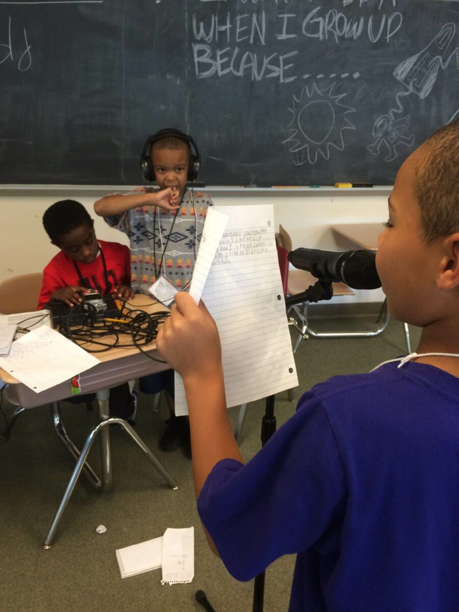 A youth holding a piece of paper and speaking into a microphone and two youths, one wearing headphones, listening to a portable recorder in a classroom