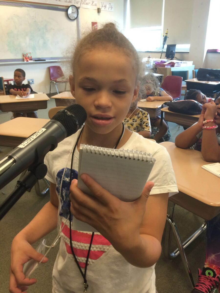 A youth looking at a notepad and speaking into a microphone in a classroom