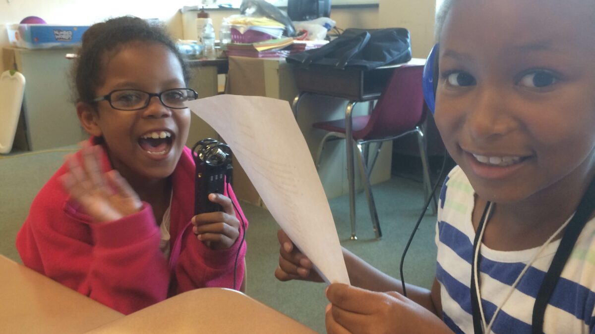 IMG_1394 A youth holding a piece of paper and a youth holding a portable recorder, both smiling at the camera in a classroom