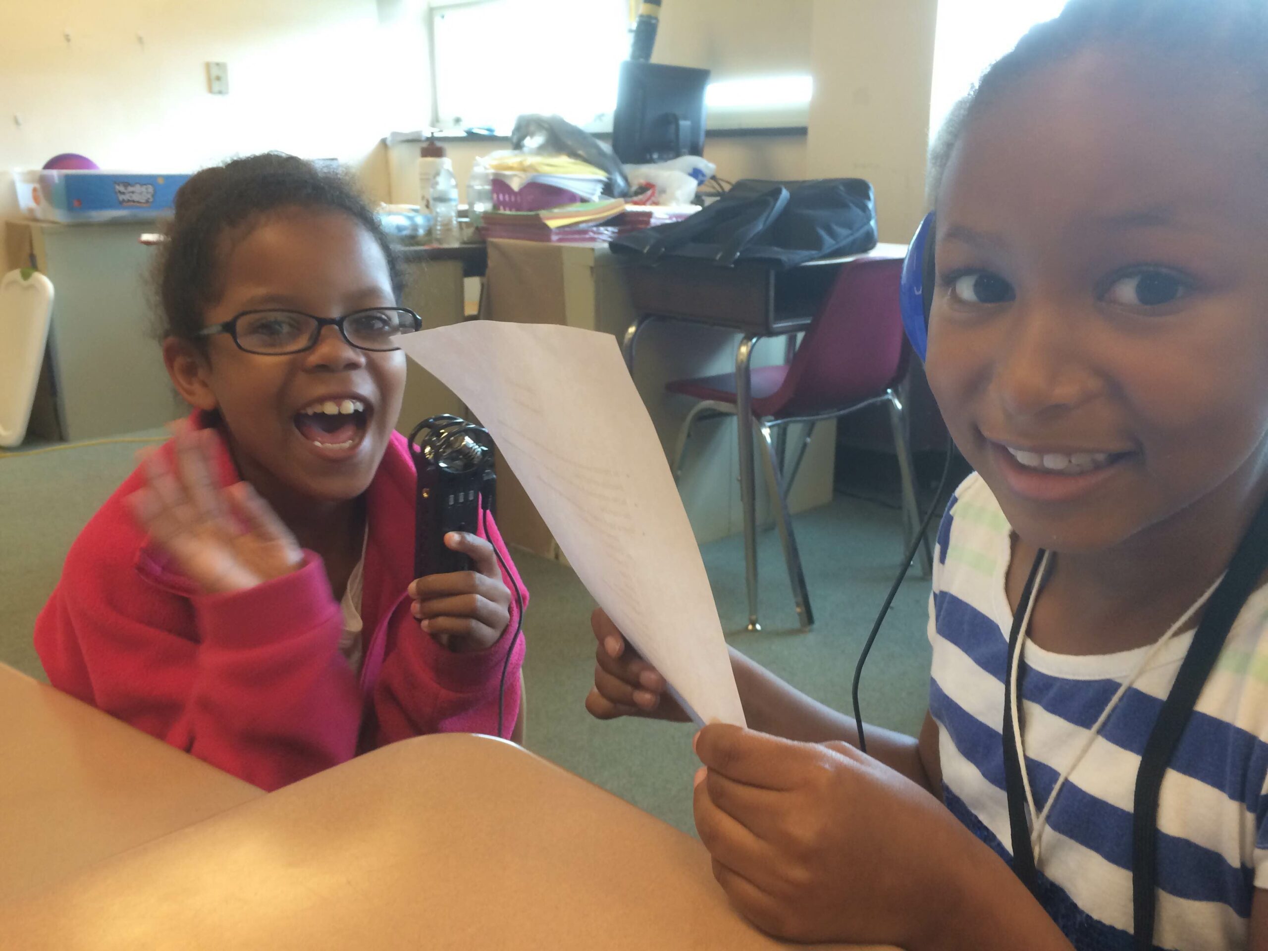 A youth holding a piece of paper and a youth holding a portable recorder, both smiling at the camera in a classroom