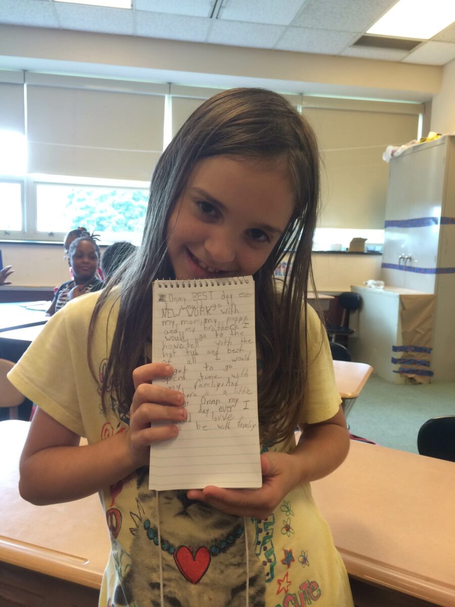 A youth holding up a notepad and smiling at the camera in a classroom