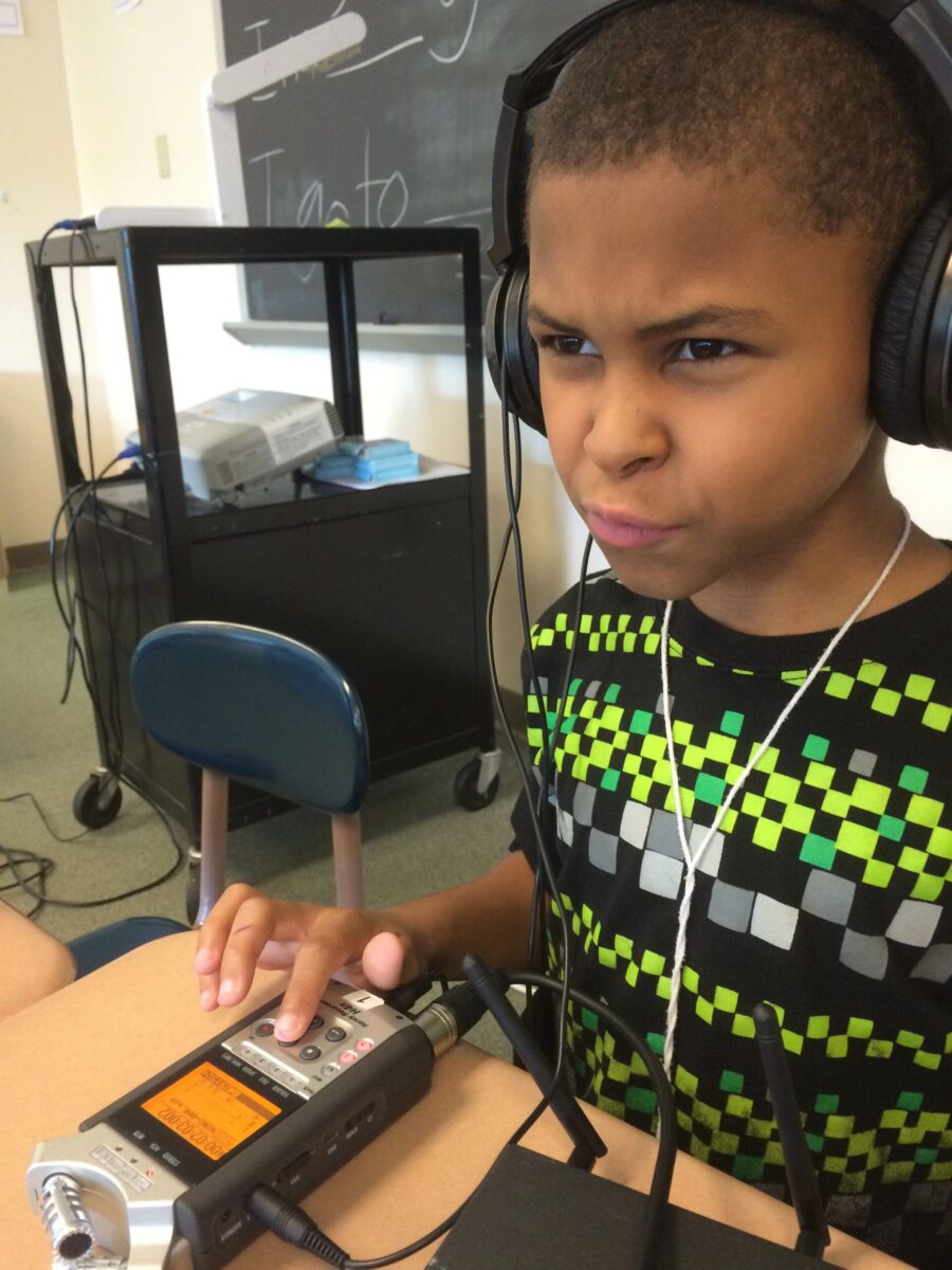 A youth wearing headphones interacting with a portable recorder on a classroom desk in front of a chalkboard