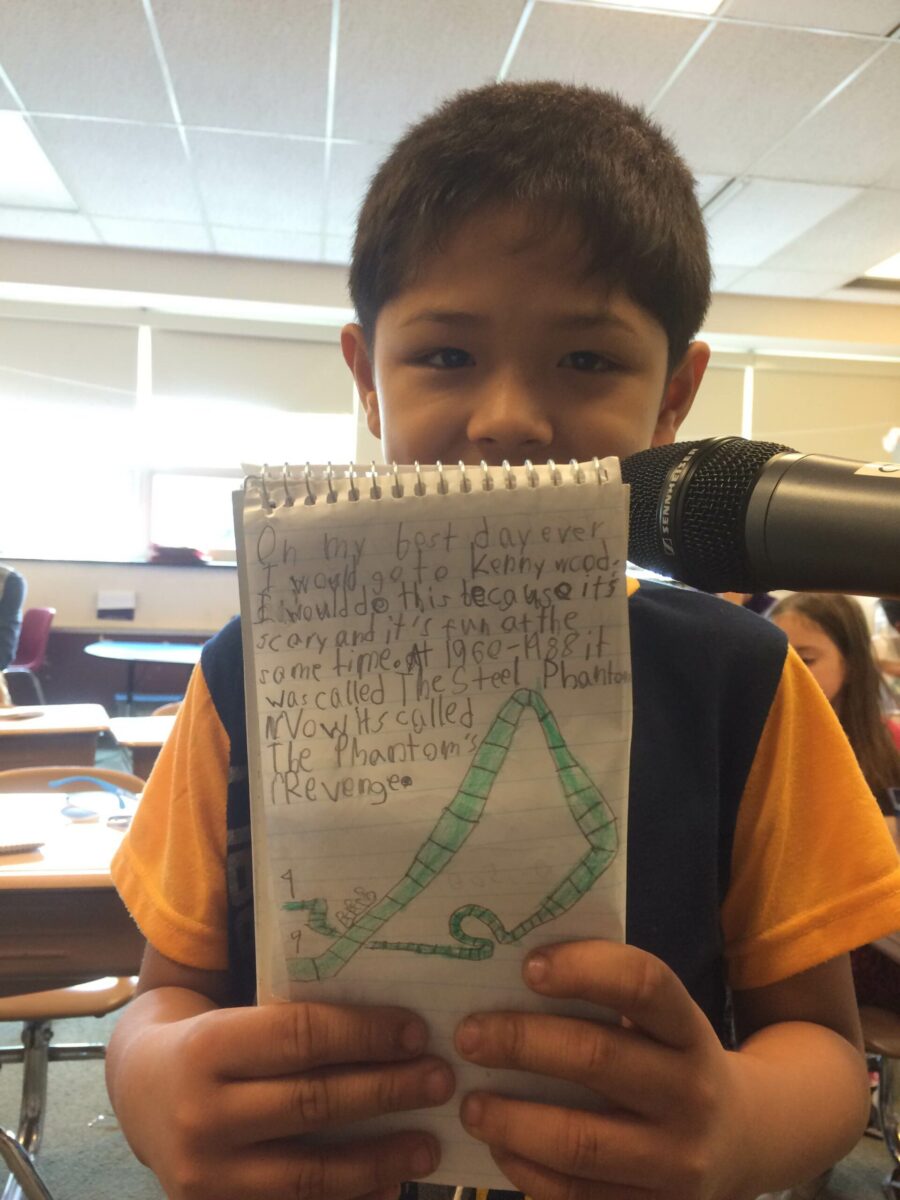 A youth holding up a notepad to the camera and standing behind a microphone in a classroom