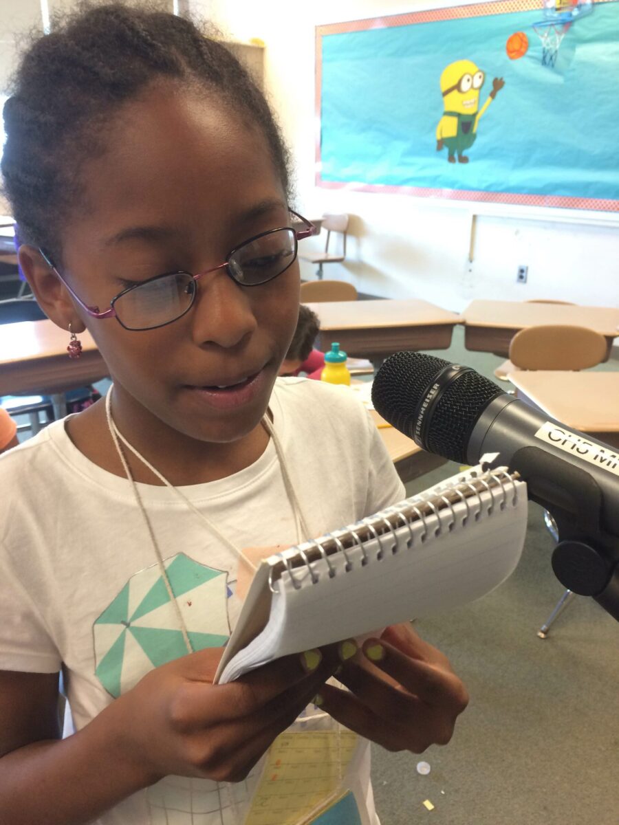 A youth holding a notepad and speaking into a microphone in a classroom