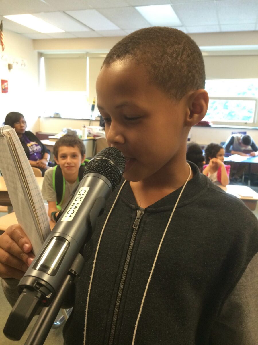 A youth holding a notepad and standing behind a microphone in a classroom