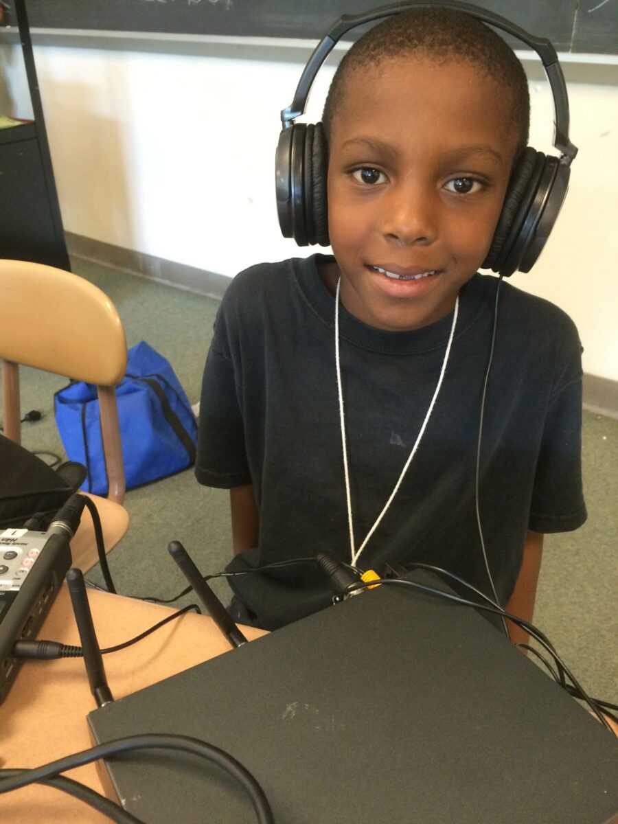 A youth wearing headphones sitting behind a portable microphone and a wireless receiver on a classroom desk