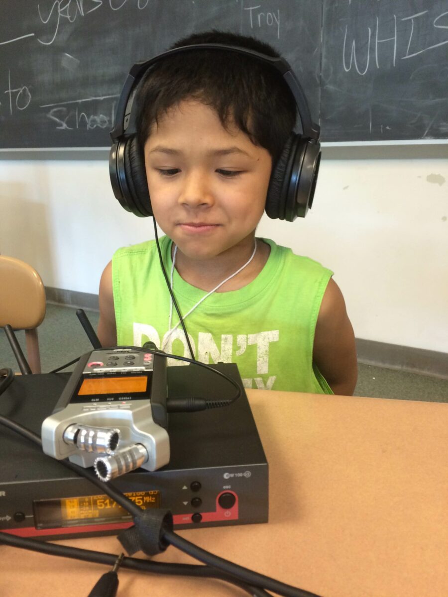 A youth wearing headphones sitting behind a portable microphone and a wireless receiver on a classroom desk