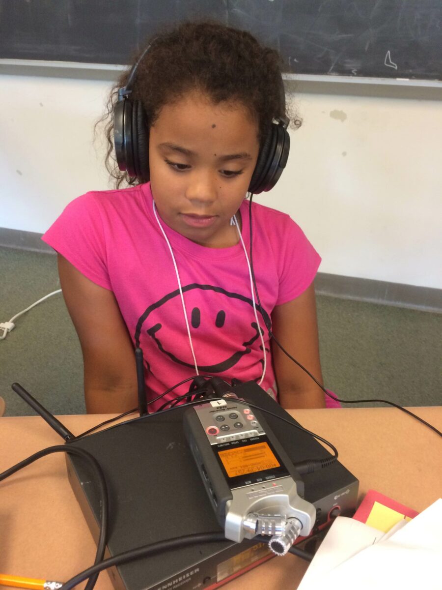A youth wearing headphones sitting behind a portable microphone and a wireless receiver on a classroom desk