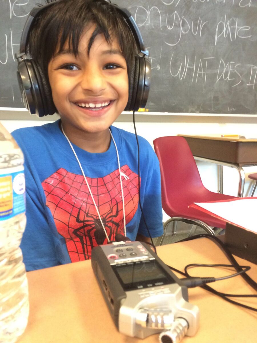 A youth wearing headphones sitting behind a portable microphone and a wireless receiver on a classroom desk