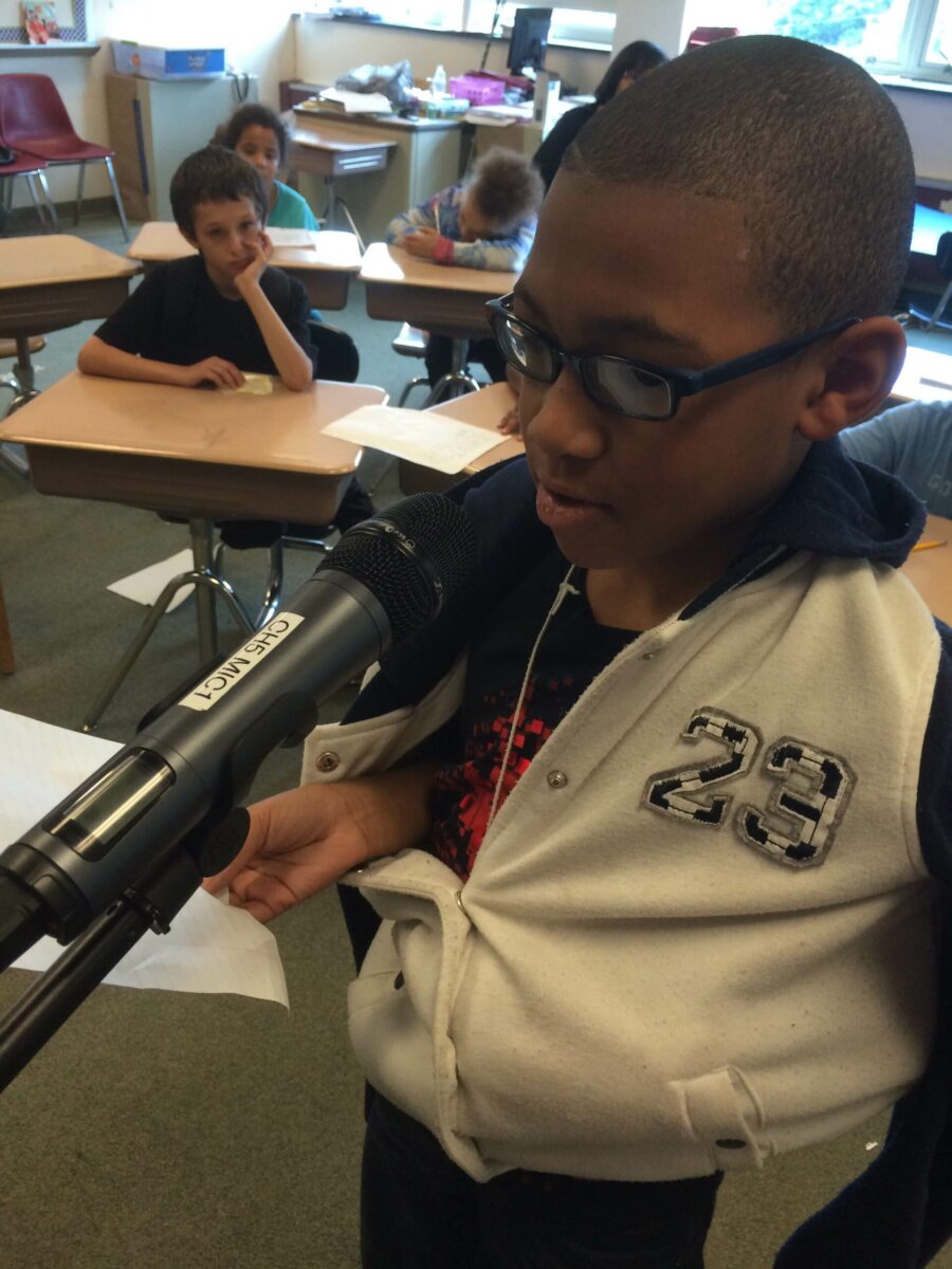 A youth holding a piece of paper and speaking into a microphone in a classroom