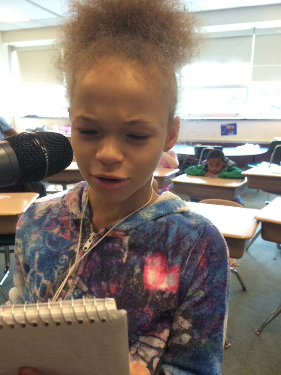 A youth holding a notepad and speaking into a microphone in a classroom