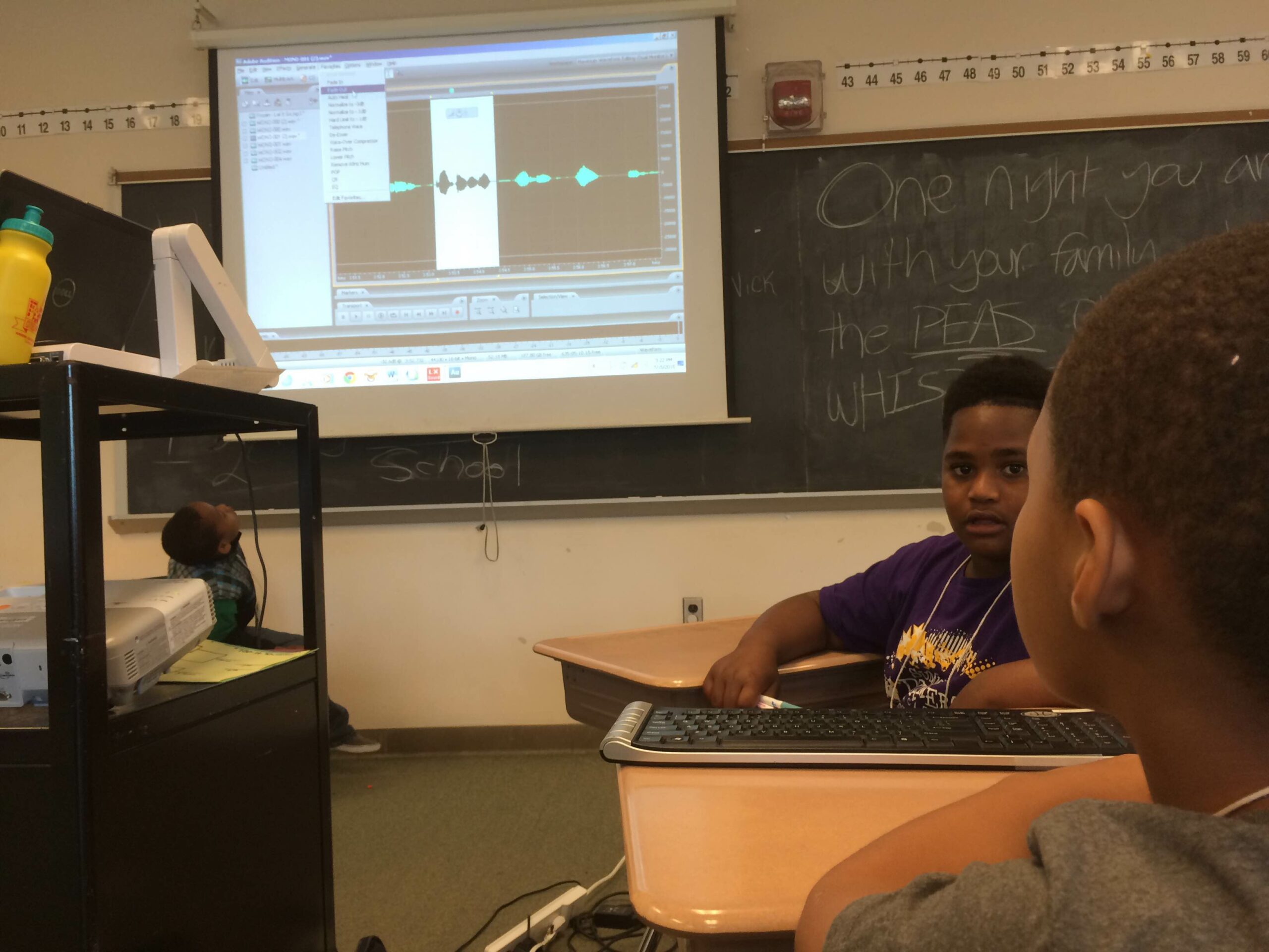 Two youth looking at audio editing software on a projected screen in front of a blackboard in a classroom