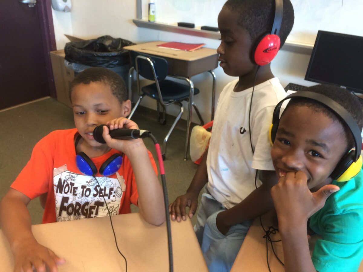 IMG_1494 Three youth wearing headphones sitting in a classroom, one holding and speaking into a microphone