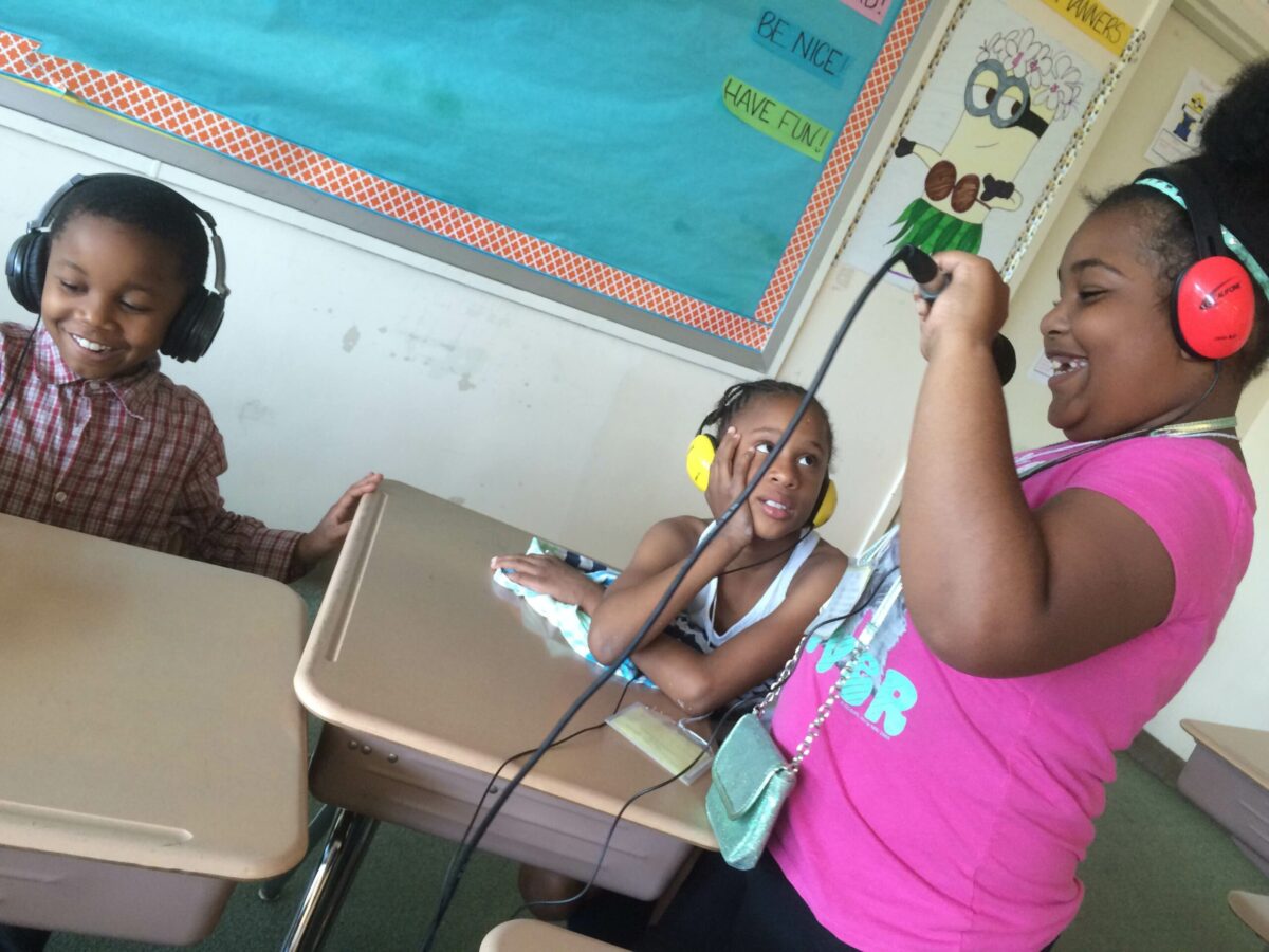 IMG_1498 Two youth wearing headphones sitting at classroom desks and one youth wearing headphones standing while holding and speaking into a microphone in a radio studio