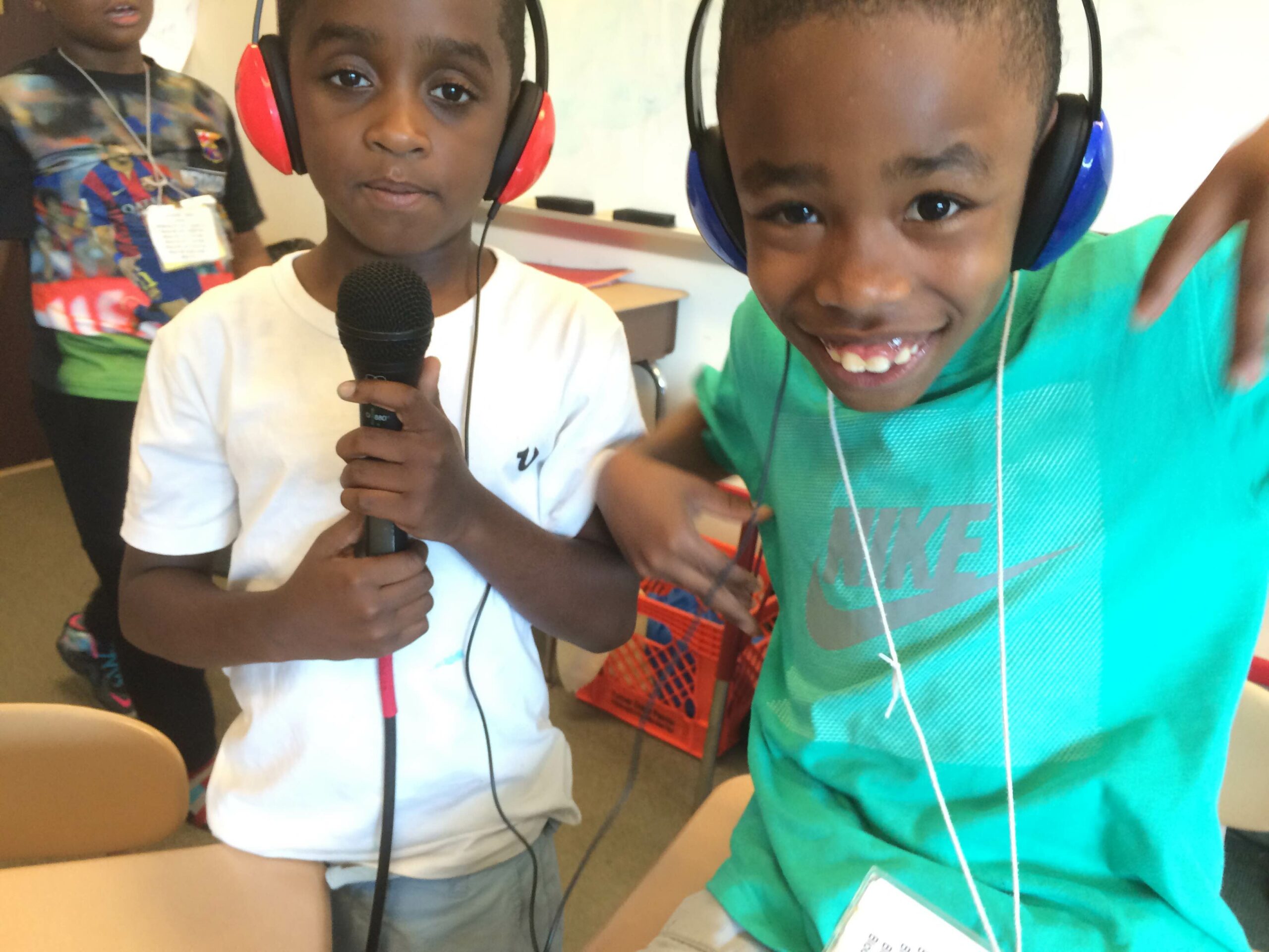 Two youth wearing headphones in a classroom, one holding a microphone, the other smiling and posing for the camera