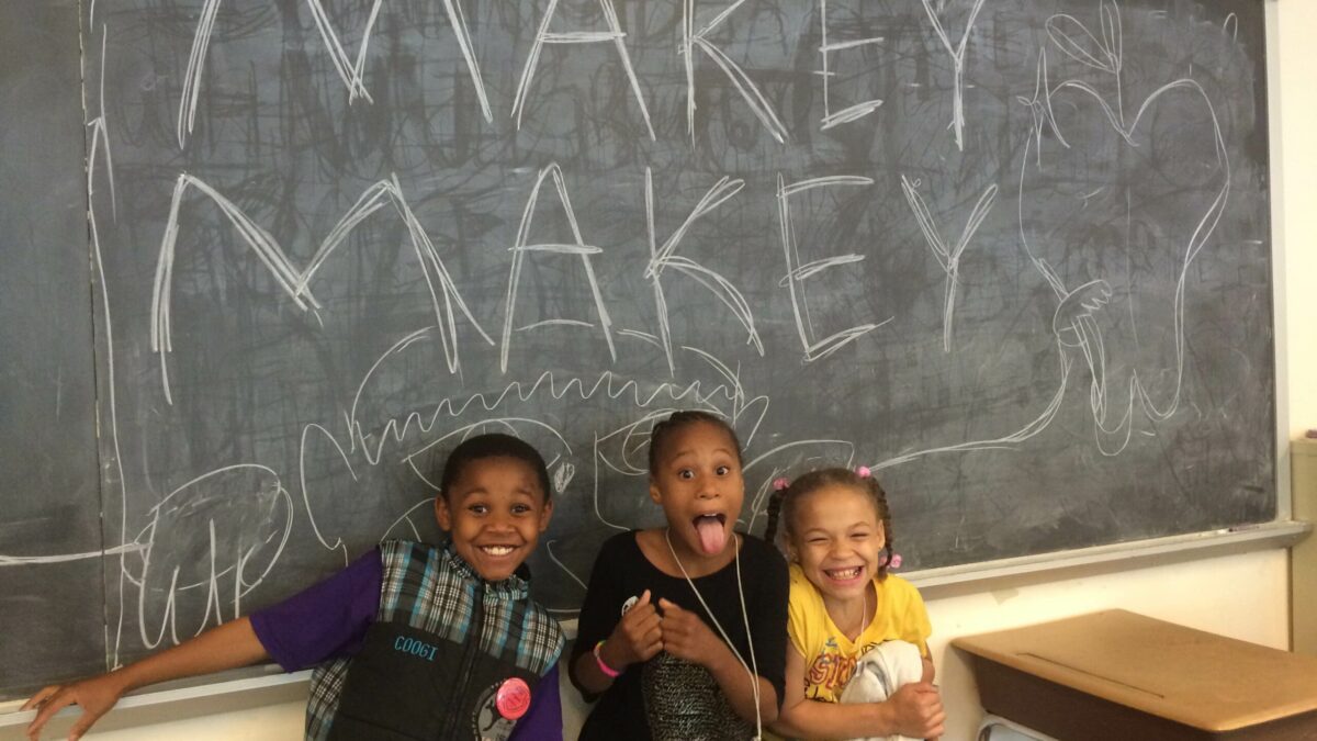 Three youth smiling and posing underneath a chalkboard with "Makey Makey" written on it