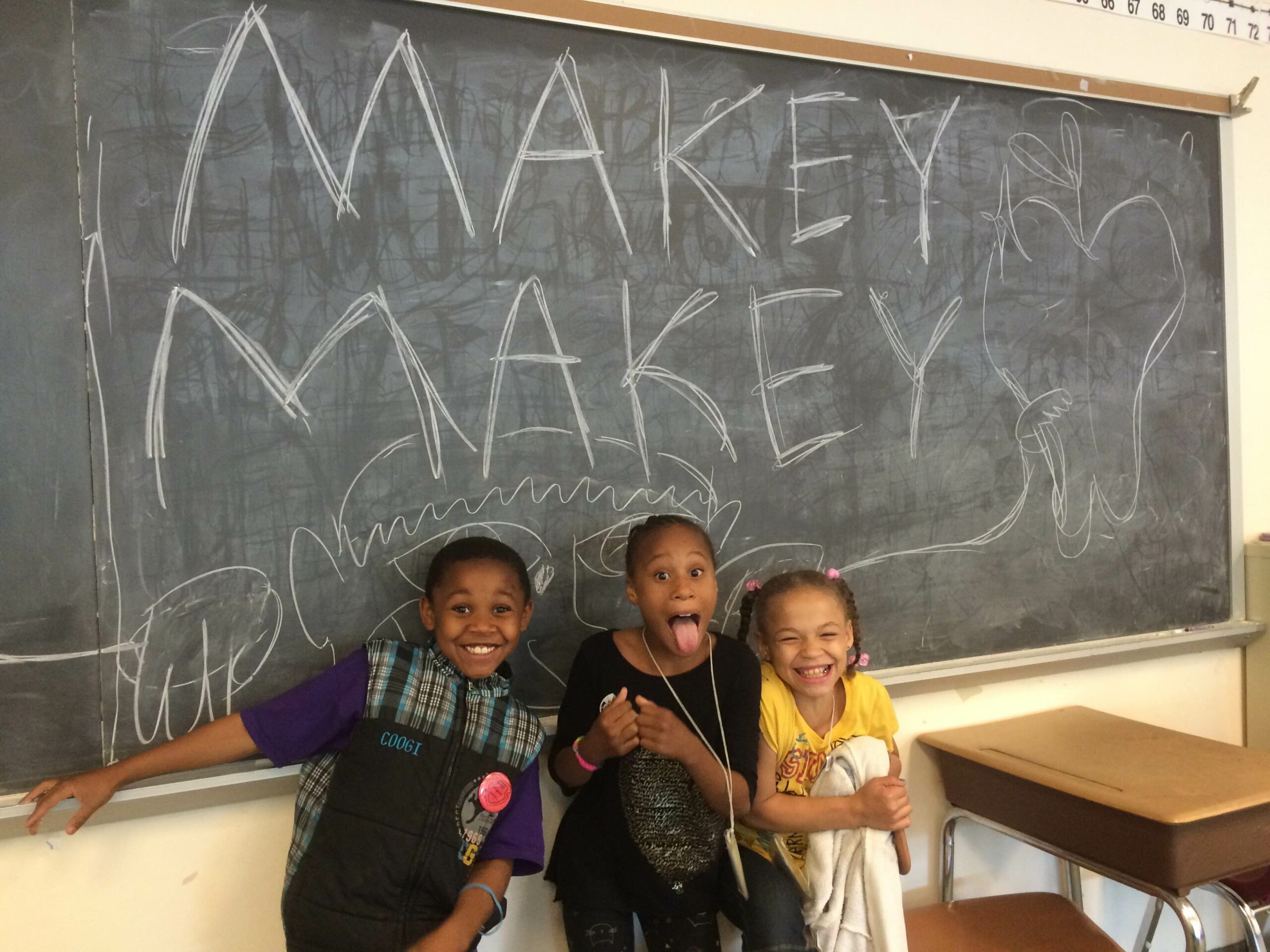 Three youth smiling and posing underneath a chalkboard with "Makey Makey" written on it
