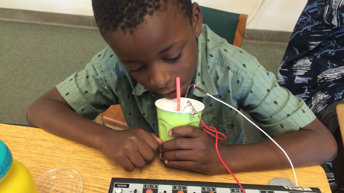 A youth speaking into a plastic cup connected to a mixer on a wooden desk