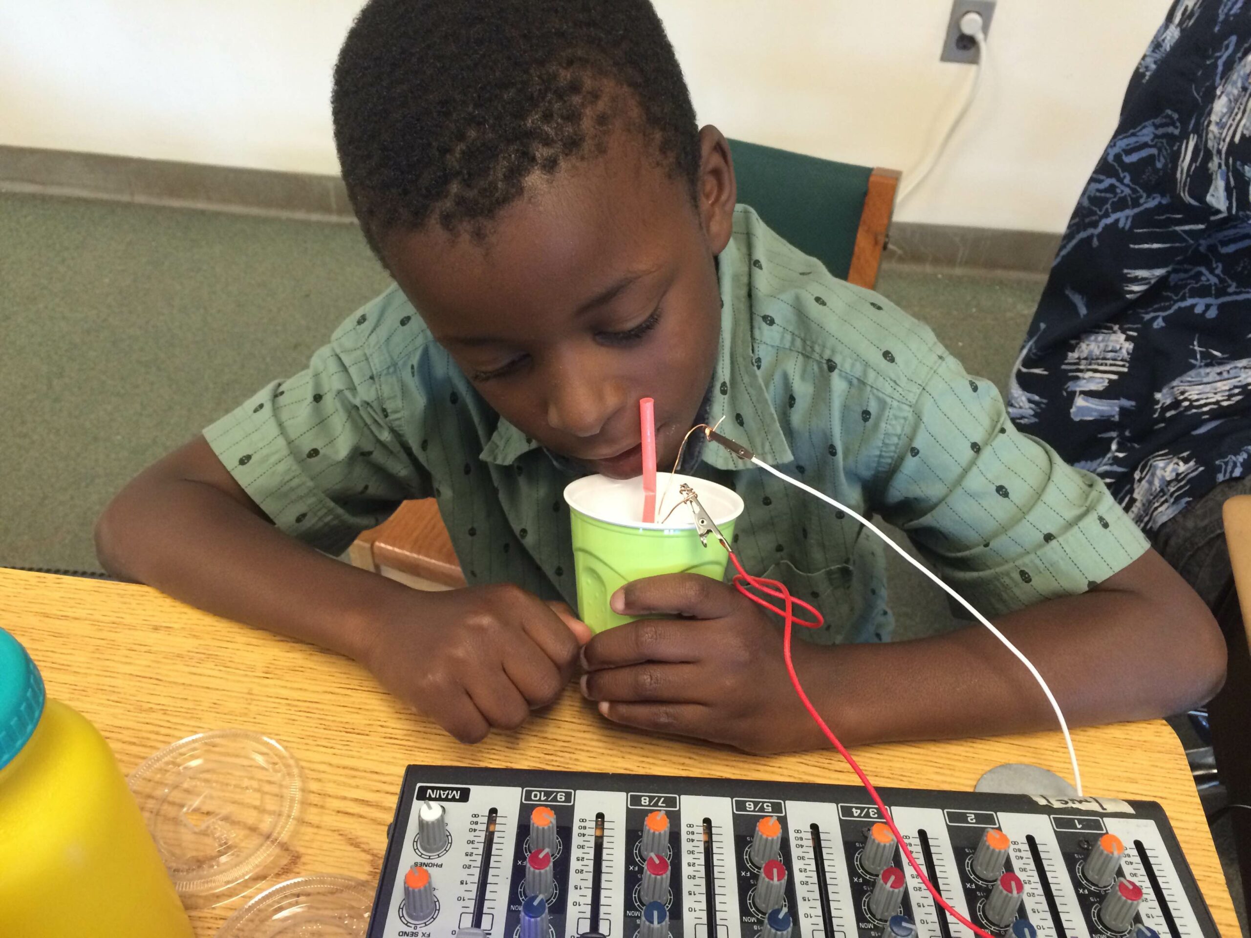 A youth speaking into a plastic cup connected to a mixer on a wooden desk