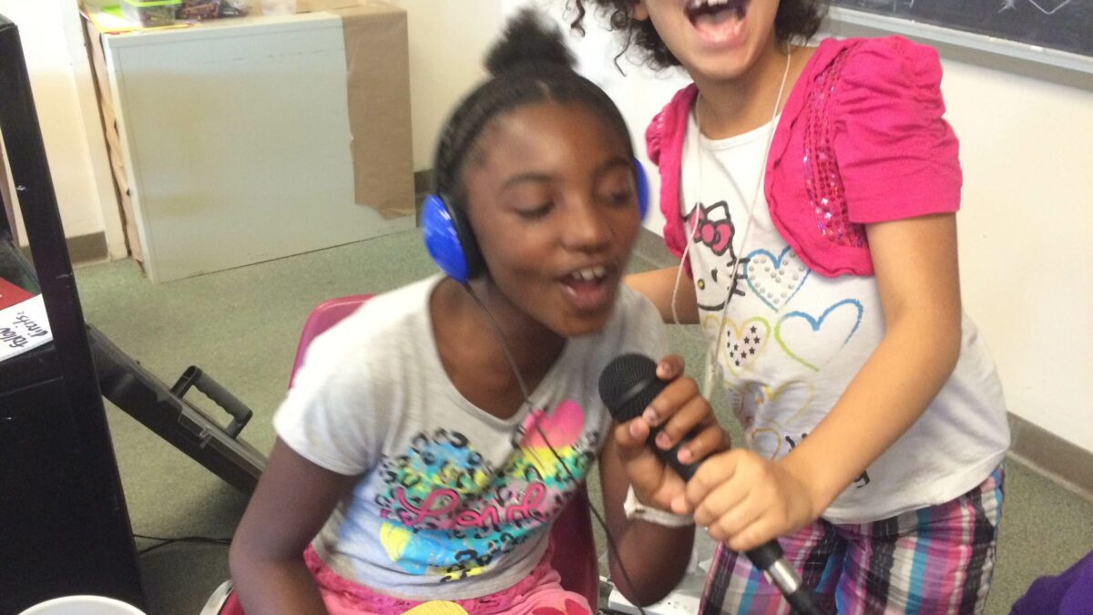 IMG_1627 Two youth holding a microphone in a classroom, one youth wearing headphones and speaking into the microphone