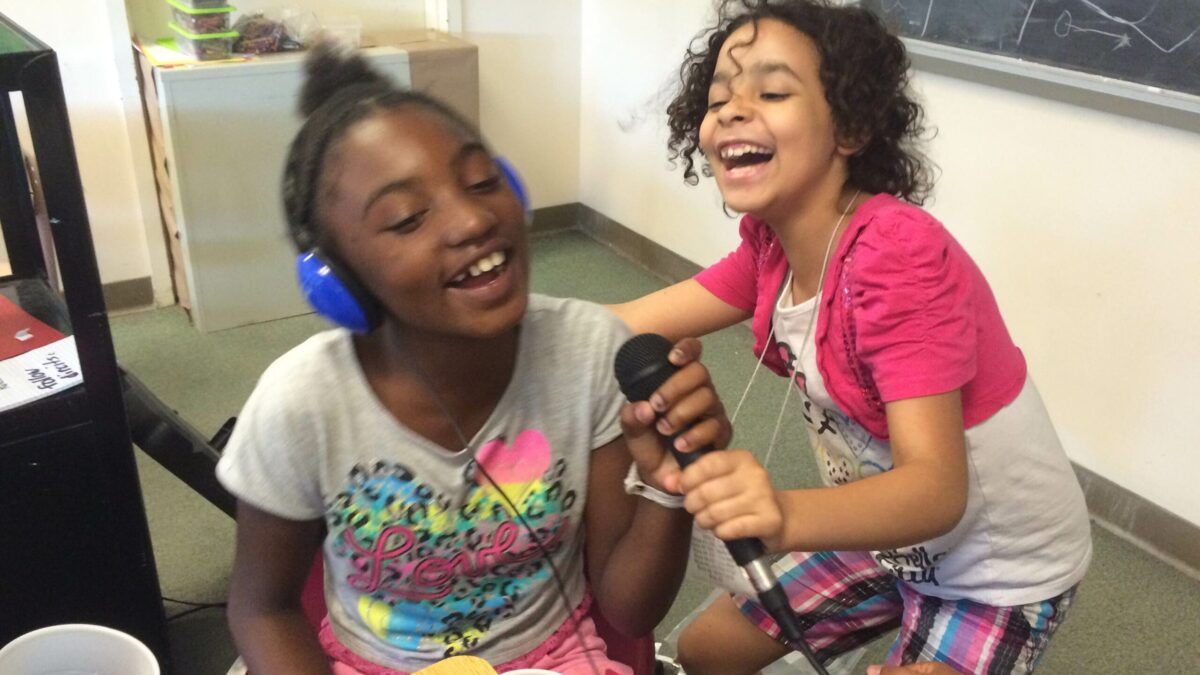 IMG_1630 Two youth holding a microphone in a classroom, one youth wearing headphones and speaking into the microphone