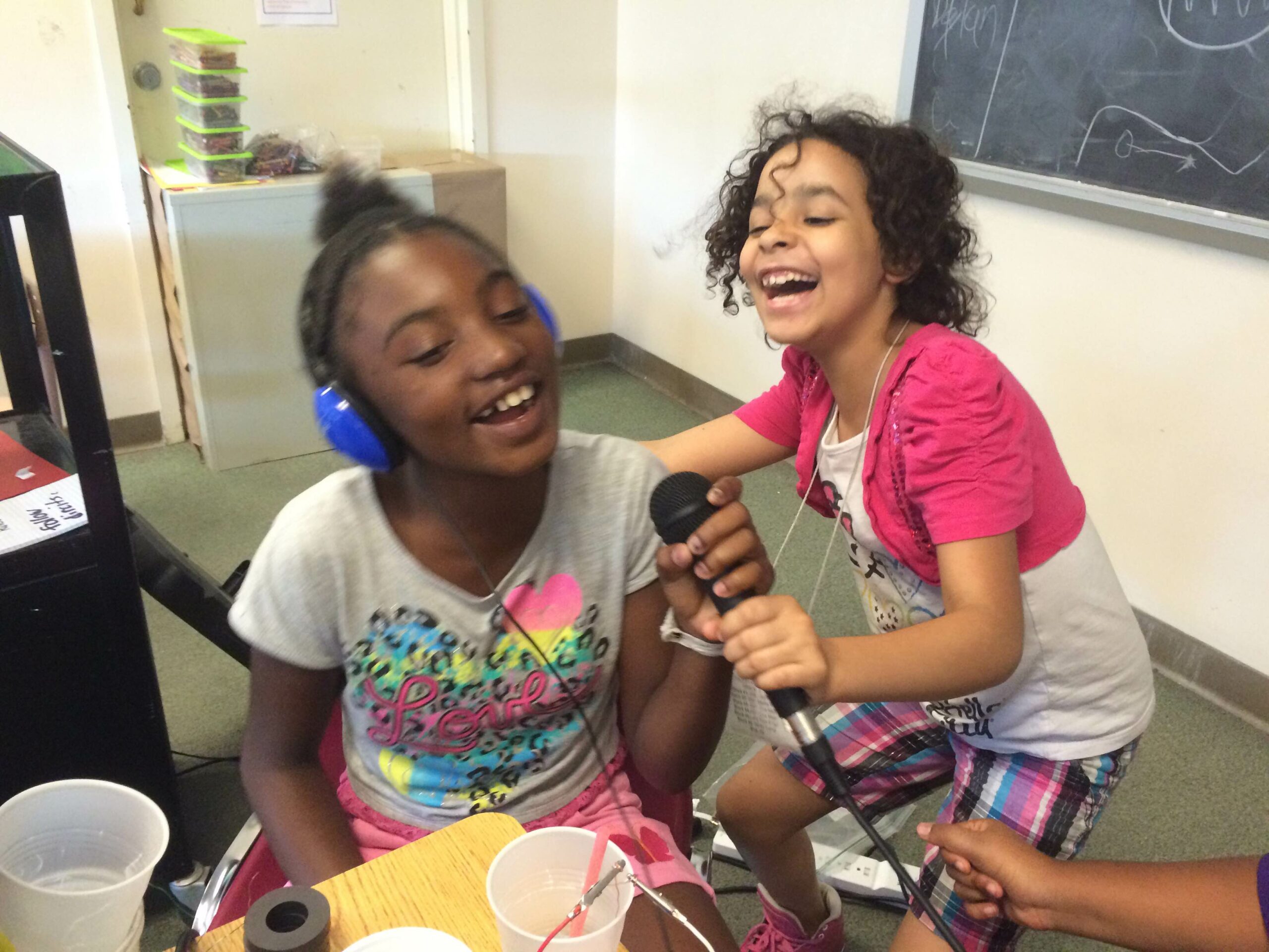 Two youth holding a microphone in a classroom, one youth wearing headphones and speaking into the microphone