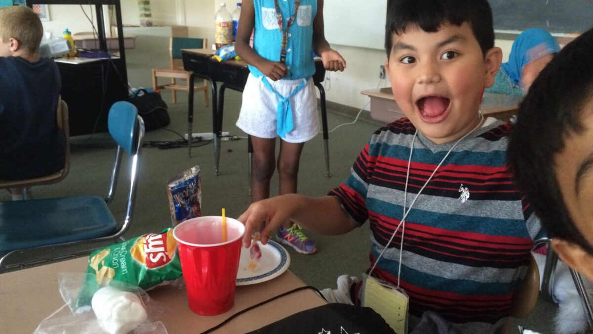 A youth with their mouth open sitting behind a red plastic cup and a bag of chips on a classroom desk