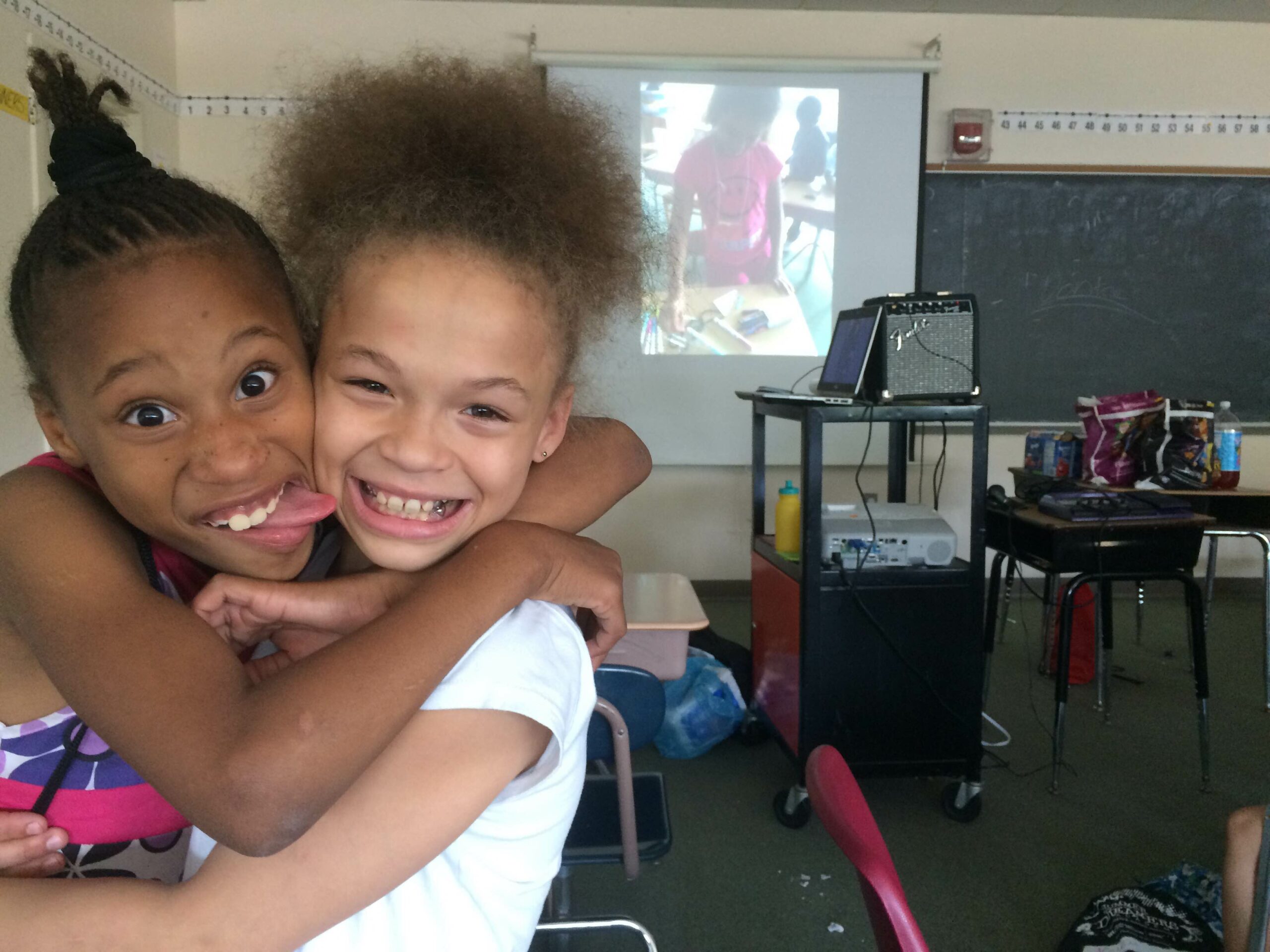 Two youth smiling in front of a projected screen in front of a chalkboard in a classroom