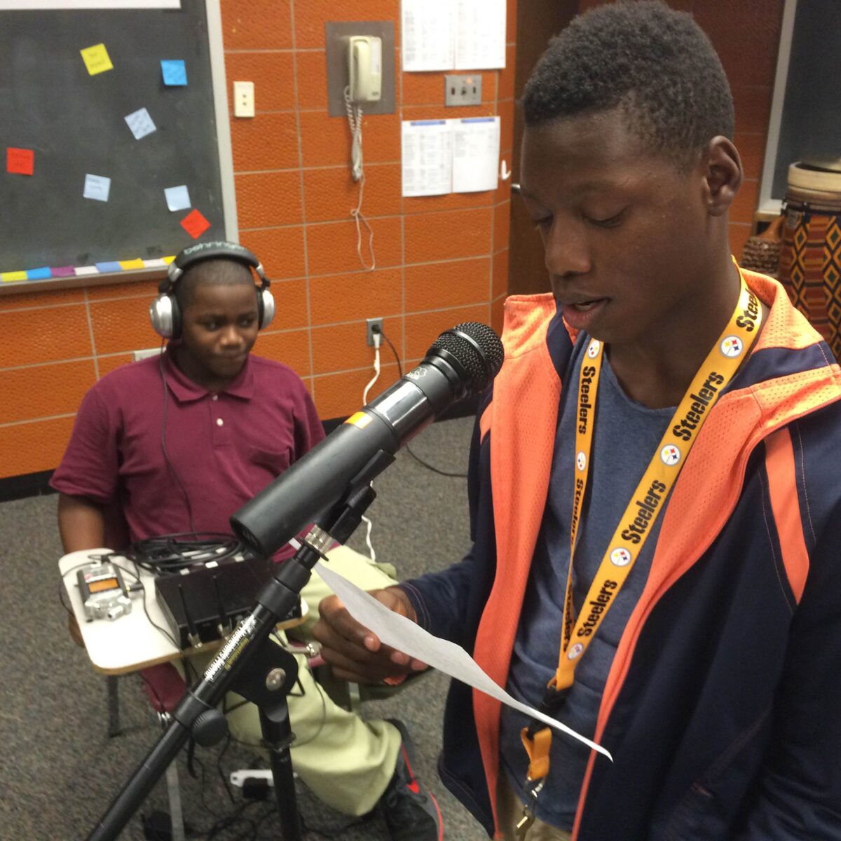 IMG_1718 A youth wearing headphones sitting behind a portable recorder on a classroom desk, and a youth reading a piece of paper into a microphone in a classroom