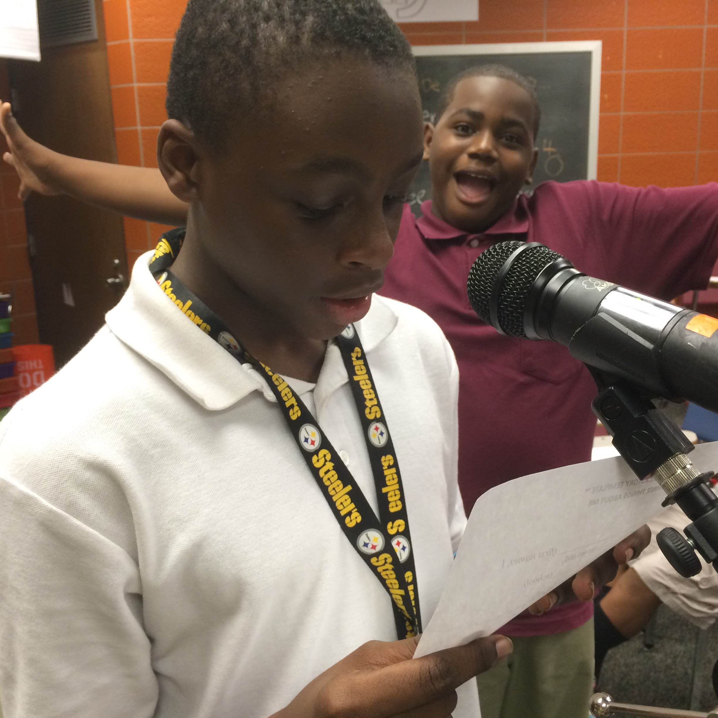 A youth reading a piece of paper into a microphone and a youth smiling and posing in the background in a classroom