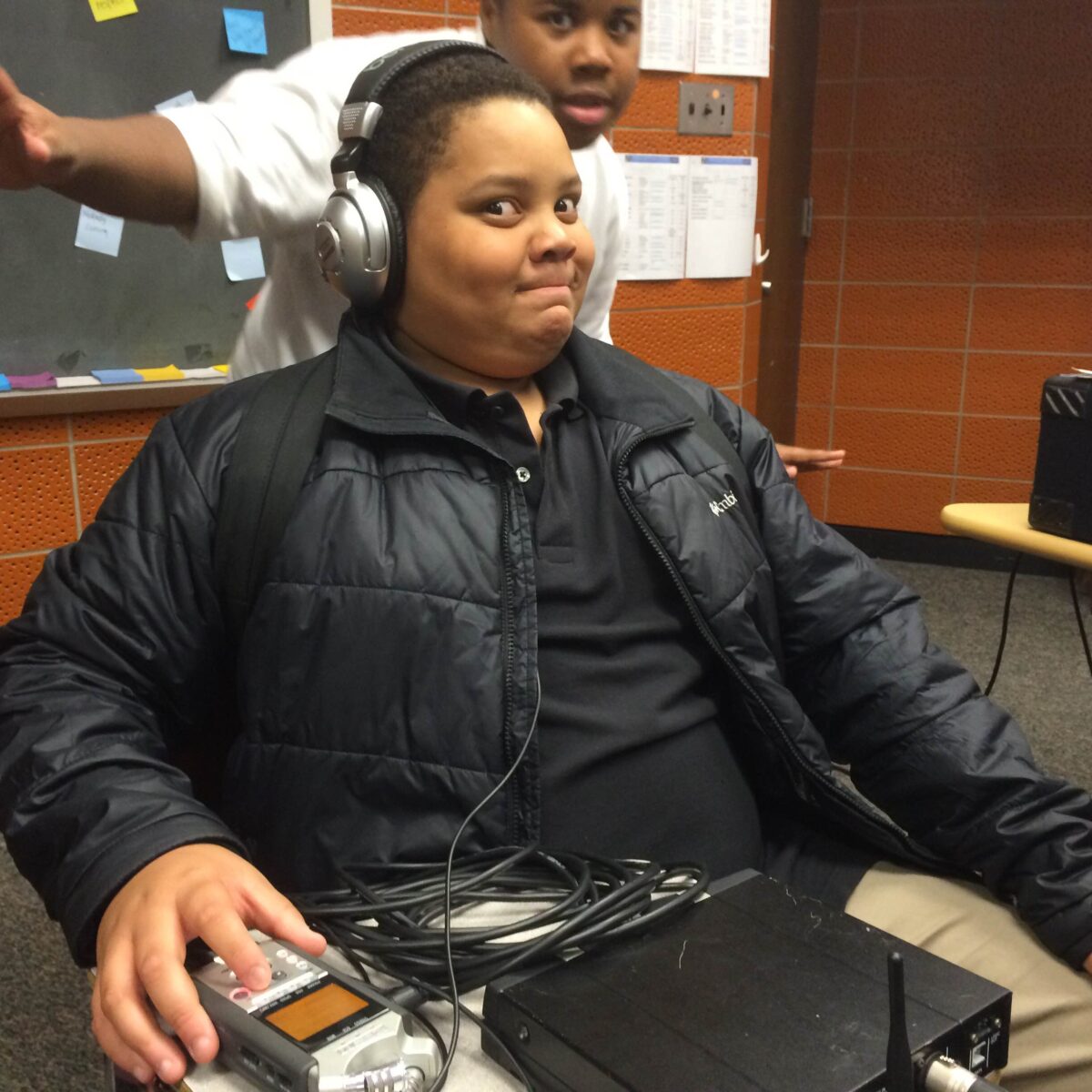 IMG_1737 A youth wearing headphones looking concerned at the camera sitting behind a portable recorder and another youth posing in the background in a classroom