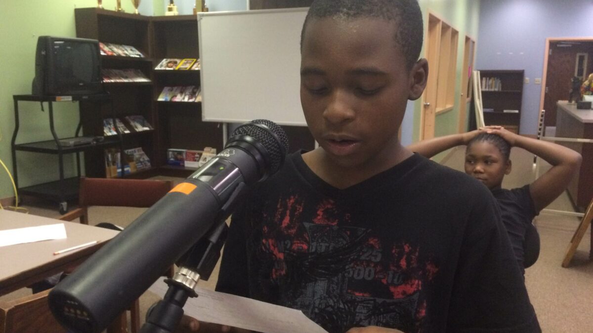 A youth holding a piece of paper speaking into a microphone in a library