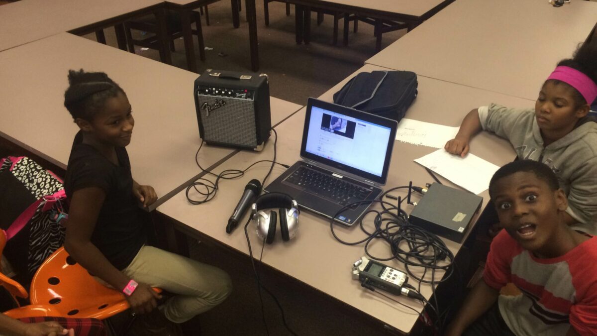Three youth sitting around audio recording equipment connected to a laptop computer on a table in a library